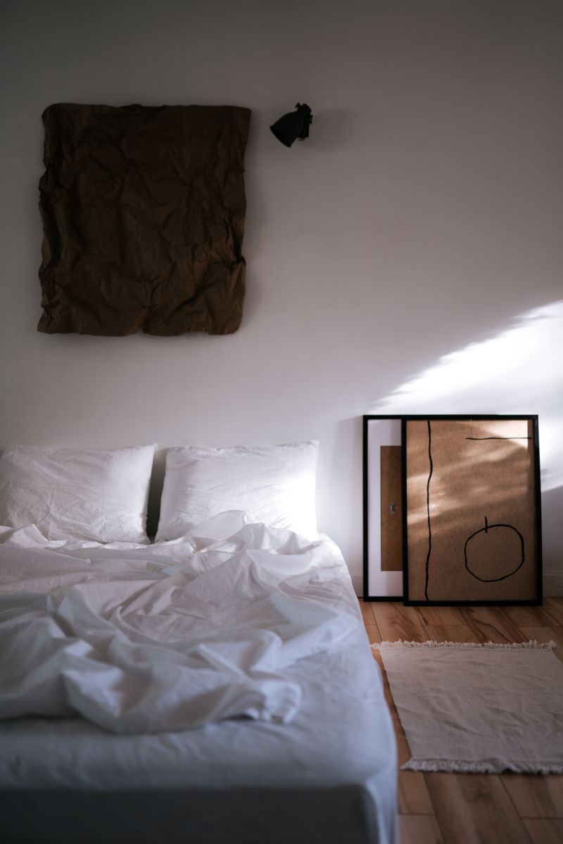 A minimalist bedroom interior featuring abstract art, linens, and natural light casting shadows.